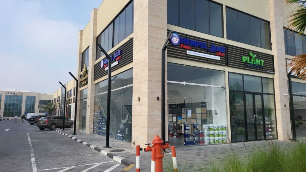 The glass storefront of a Hempel Paints store, featuring displays of exterior paint and stacks of paint for paint wholesale dealers. It serves as a resource for painting contractors near me and customers seeking paint suppliers near me.