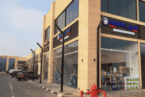 The glass storefront of a Al-Khabeer paints store, featuring displays of exterior paint and stacks of paint for paint wholesale dealers. It serves as a resource for painting contractors and customers seeking paint suppliers.