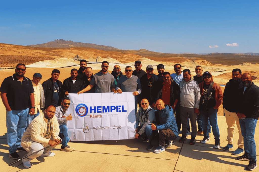 A large group of Hempel Paints Achievers Club members posing with a company banner in a desert landscape.